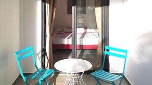 A balcony with two blue chairs, a white table, and a bed with red bedding in the background. - Seasonal rental brand new - 4 pers. -POSSESSION-end construction Oct. 19 (LA POSSESSION)