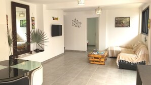 A living room with a sofa, a glass coffee table, a TV, and a dining area with a table and chairs. - Seasonal rental brand new - 4 pers. -POSSESSION-end construction Oct. 19 (LA POSSESSION)