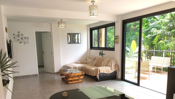 A modern living room with a sofa, a coffee table, and a dining table. There is a large window with a view of greenery outside. - Seasonal rental brand new - 4 pers. -POSSESSION-end construction Oct. 19 (LA POSSESSION)