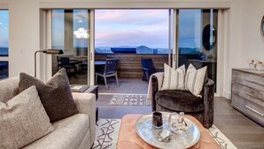 Een moderne woonkamer met een bank, fauteuil en salontafel. Grote glazen deuren geven uitzicht op een balkon met uitzicht op de bergen.