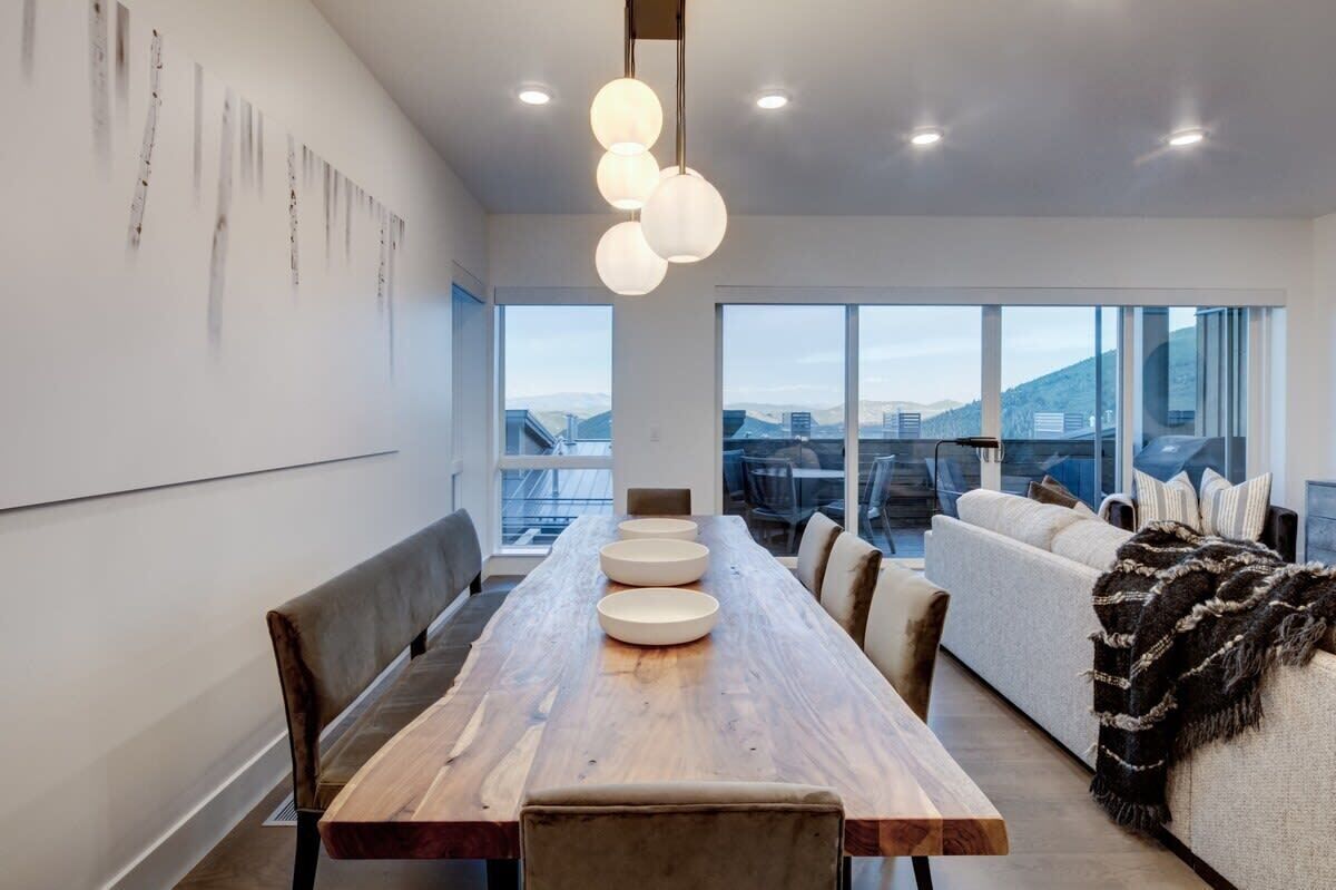 A modern dining area with a long wooden table, multiple chairs, and a large window offering a view of the cityscape.