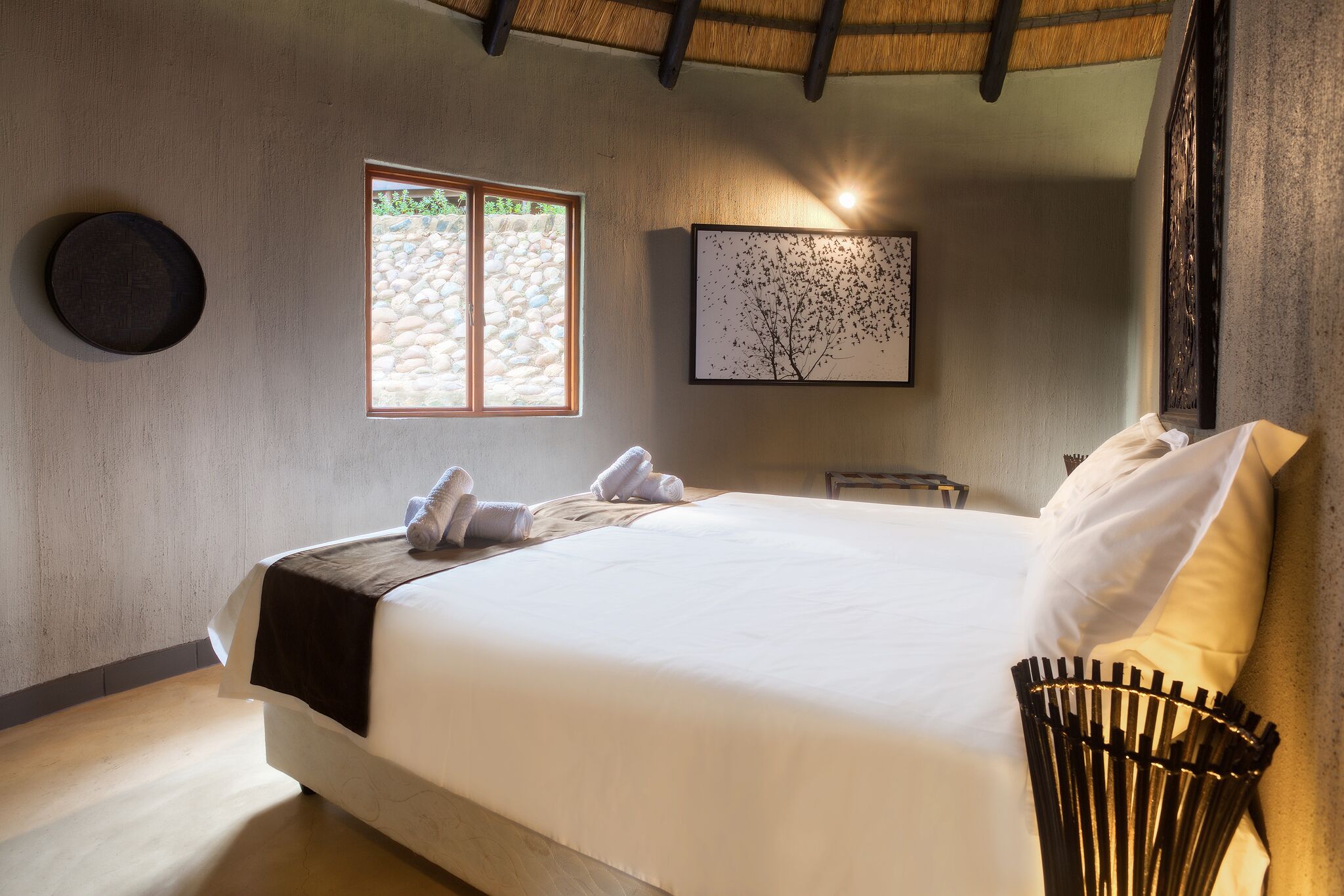 A neatly made bed with white linens and a black throw blanket, a round wall clock, a framed artwork, and a window with a stone wall view.