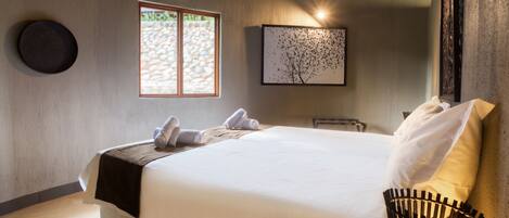 A neatly made bed with white linens and a black throw blanket, a round wall clock, a framed artwork, and a window with a stone wall view.