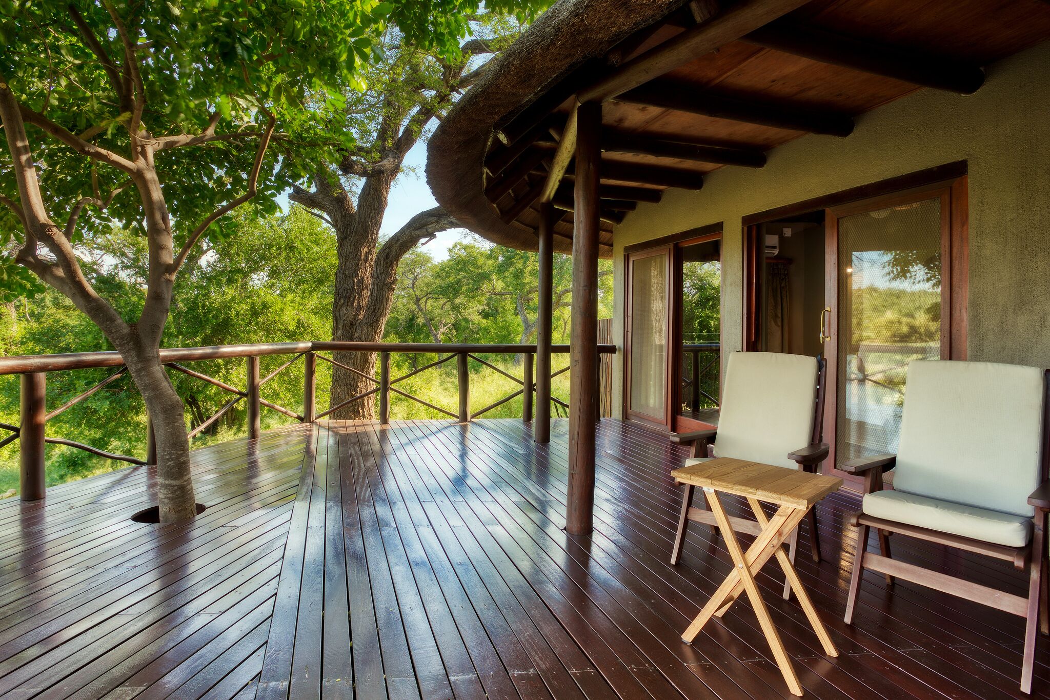 A wooden deck with a thatched roof, two white cushioned chairs, and a wooden table.