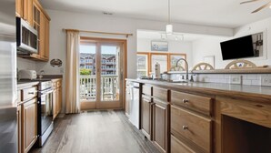 Eine Küche mit Holzausstattung, einem Edelstahlkühlschrank, einem Einbauofen, einer großen Kochinsel mit Spüle und Blick durch die Glastür nach draußen.
