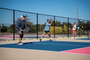 A tennis court with three players, a chain-link fence, and a clear sky.