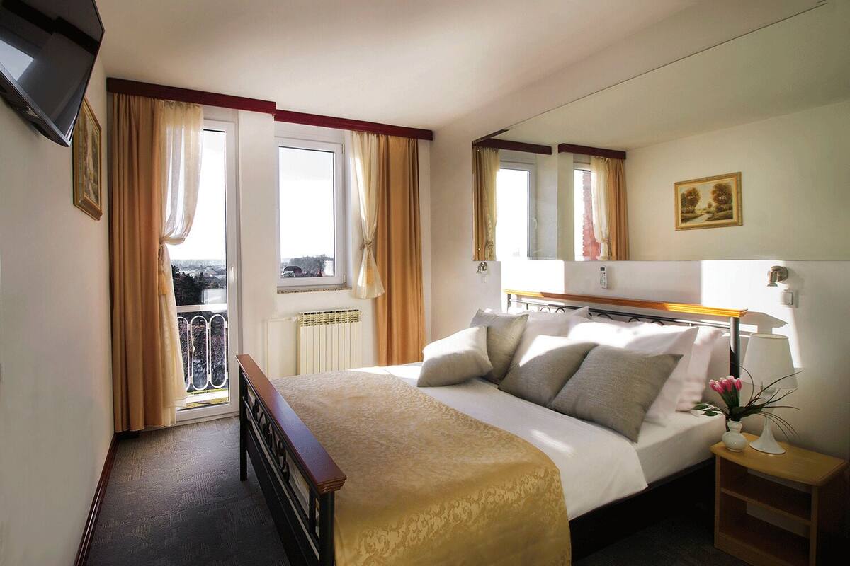 A hotel room with a bed, bedside table, lamp, and a view of the outside.
