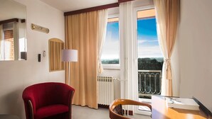 A hotel room with a red armchair, a wooden chair, a table, a lamp, and a view of the outdoors through large windows. - Hotel Slavonija (Vinkovci)