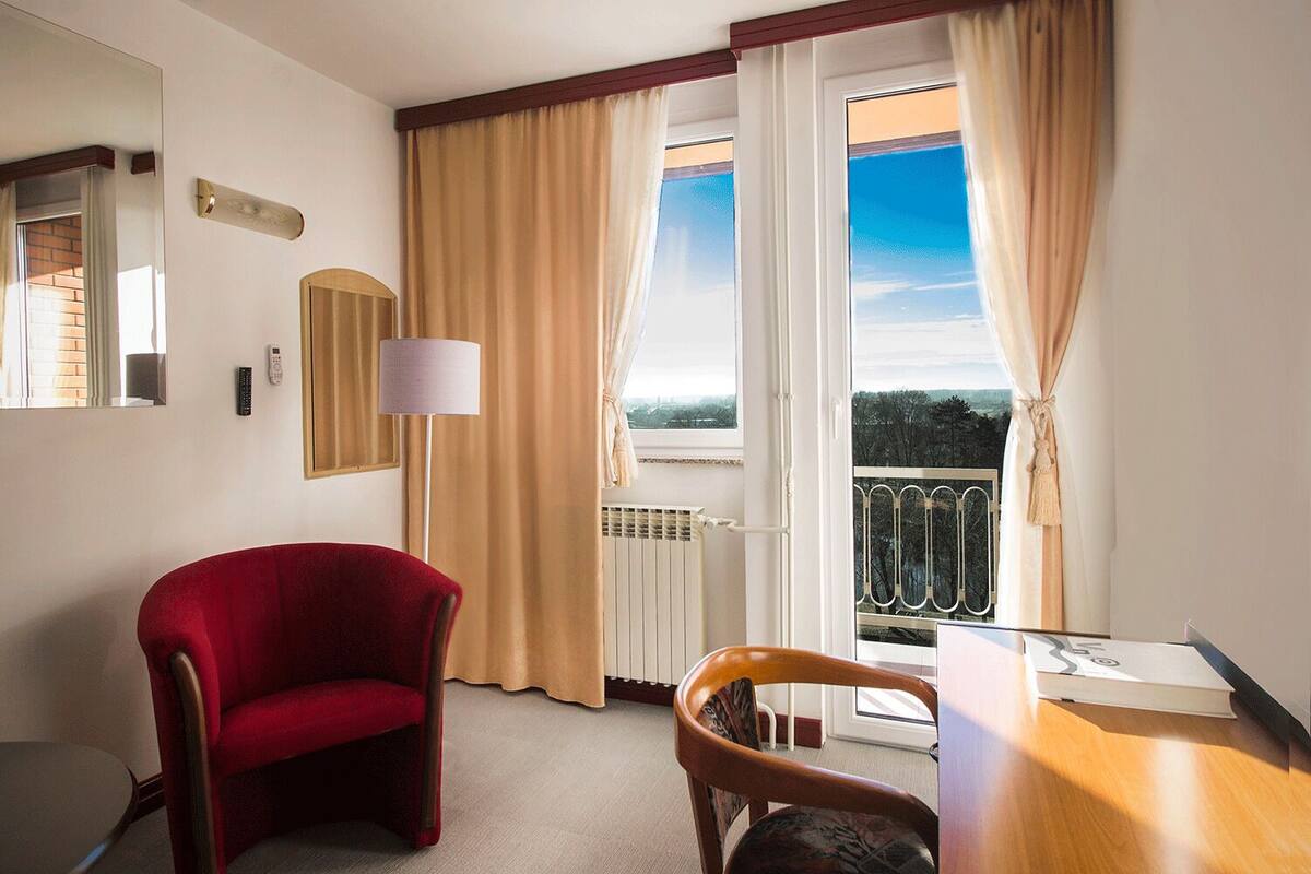 A hotel room with a red armchair, a wooden chair, a table, a lamp, and a view of the outdoors through large windows.