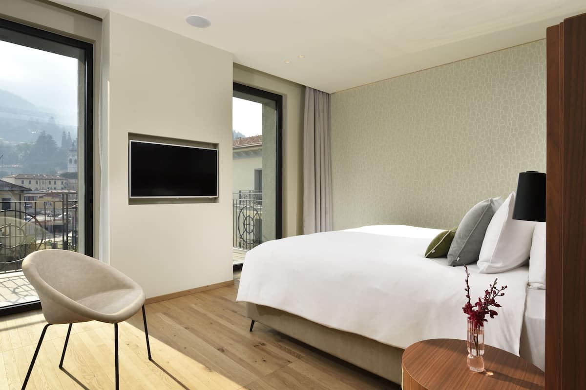 A modern hotel room with a large bed, a flat-screen TV, a chair, and a view of the city through a large window.