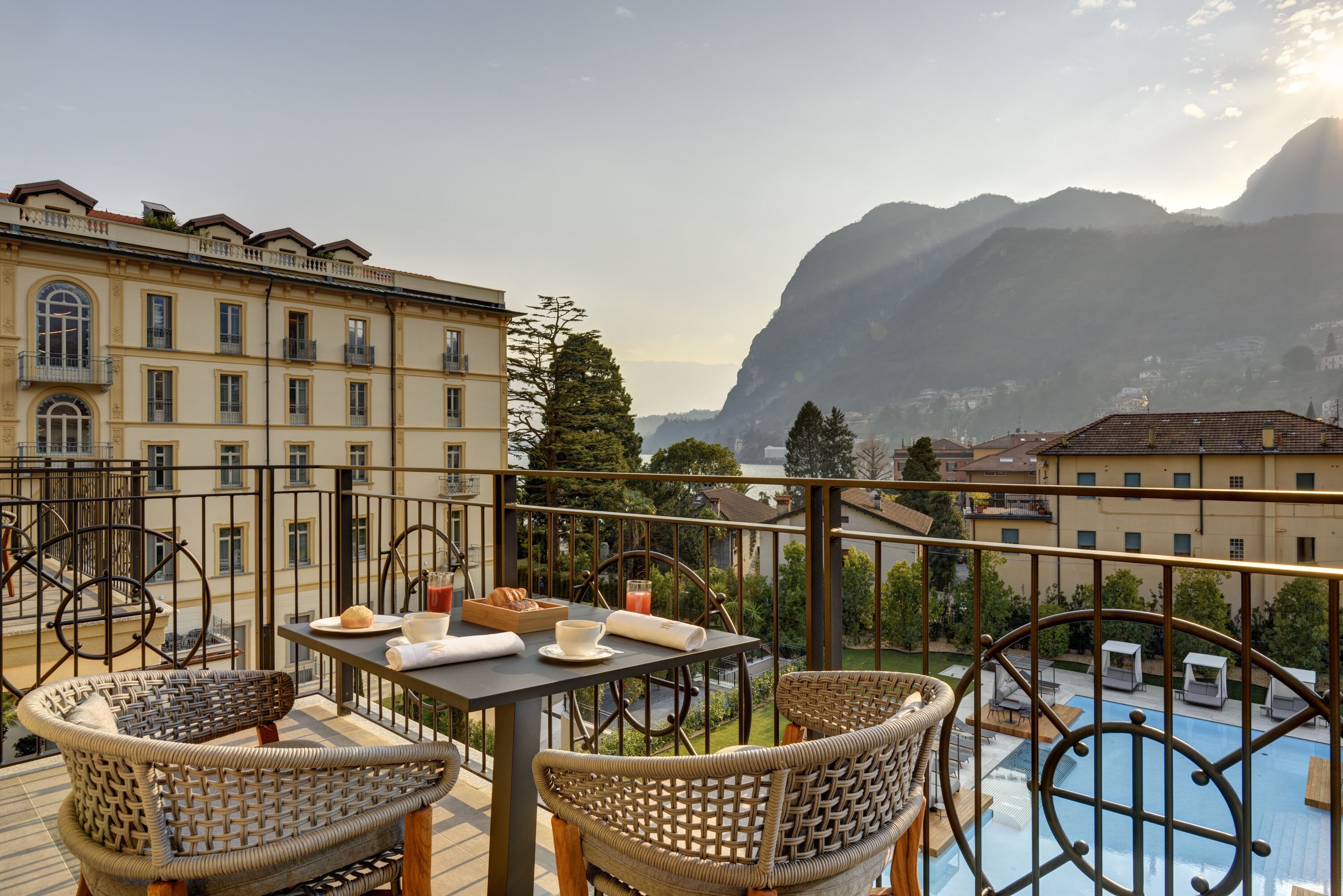 A balcony with wicker furniture, a table with breakfast items, and a view of mountains and a swimming pool.