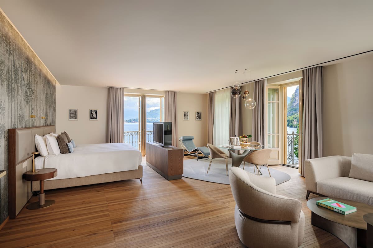 A modern hotel room with a bed, a desk, a TV, a balcony, and a seating area.