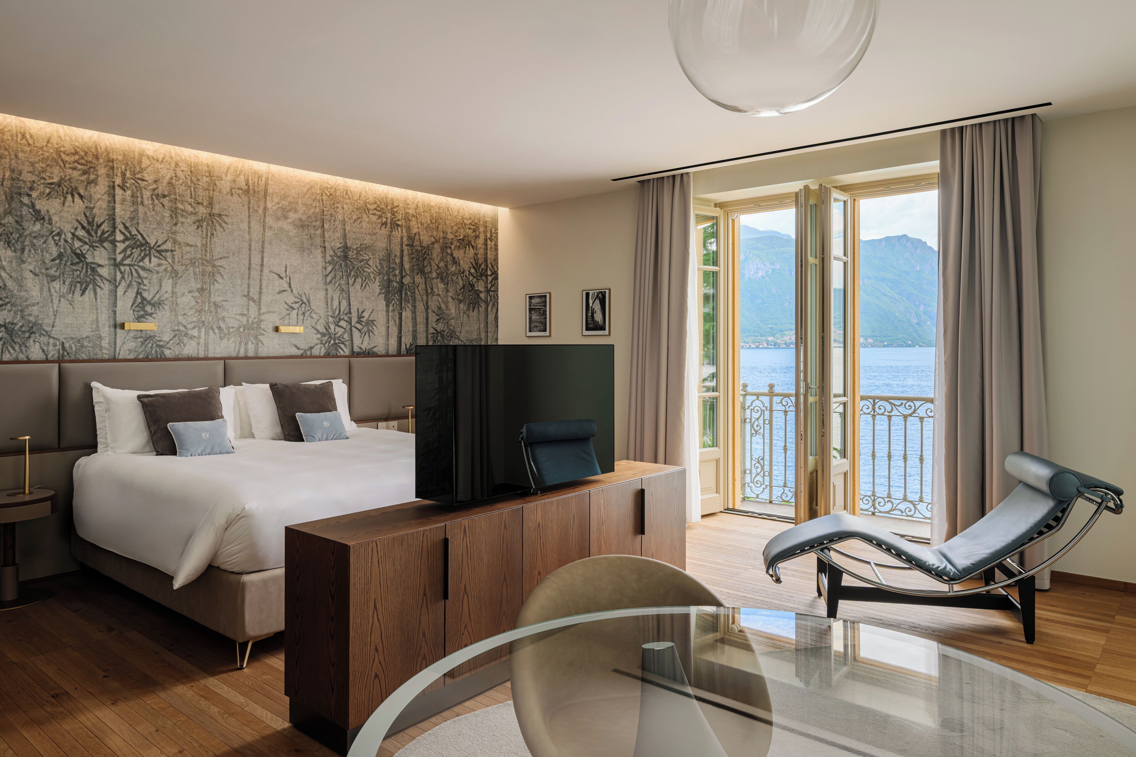A modern hotel room with a large bed, a TV, a glass table, a lounge chair, and a balcony with a view of the sea.