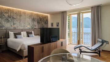 A modern hotel room with a large bed, a TV, a glass table, a lounge chair, and a balcony with a view of the sea.