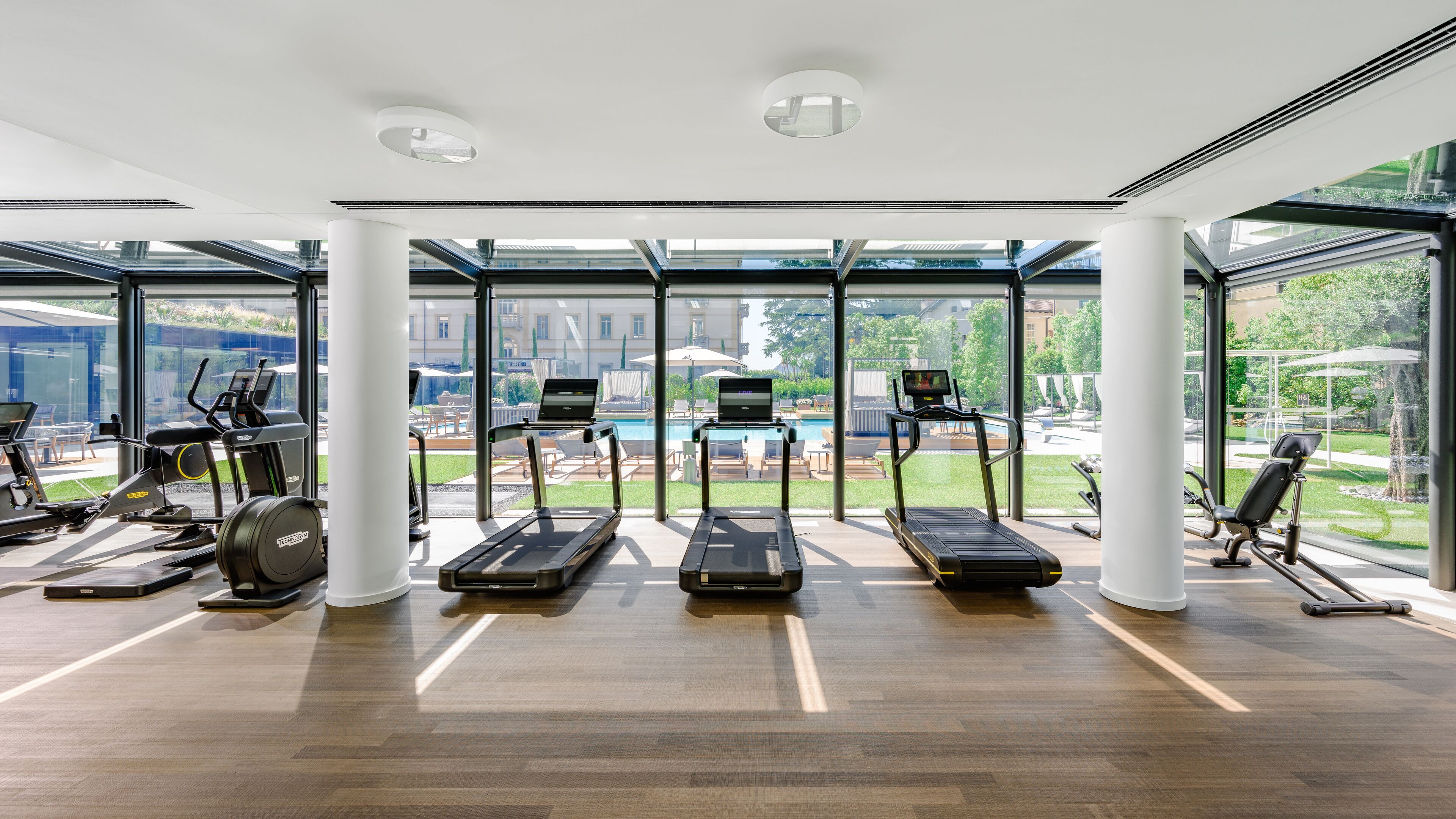 Un gimnasio con cintas de correr, elípticas y una zona de piscina visibles a través de grandes ventanales.