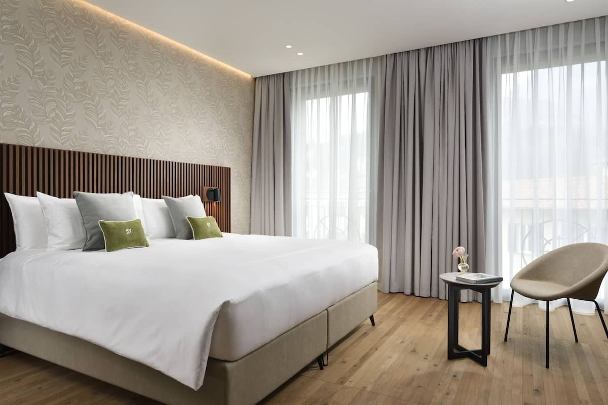 A modern hotel room with a large bed, a small table, a chair, and a view of the city through large windows.