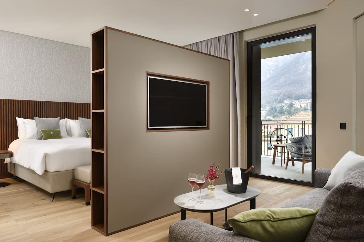 A modern hotel room with a bed, a small table with drinks, a sofa, a TV, and a balcony with a view of the mountains.