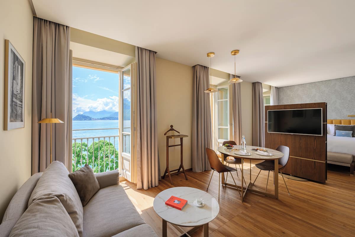 A modern hotel room with a large window offering a scenic view, a cozy seating area, a dining table, and a flat-screen TV.