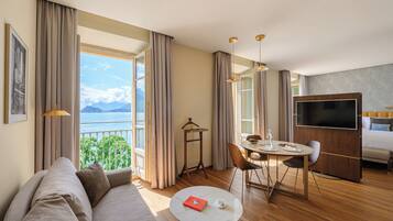 A modern hotel room with a large window offering a scenic view, a cozy seating area, a dining table, and a flat-screen TV.