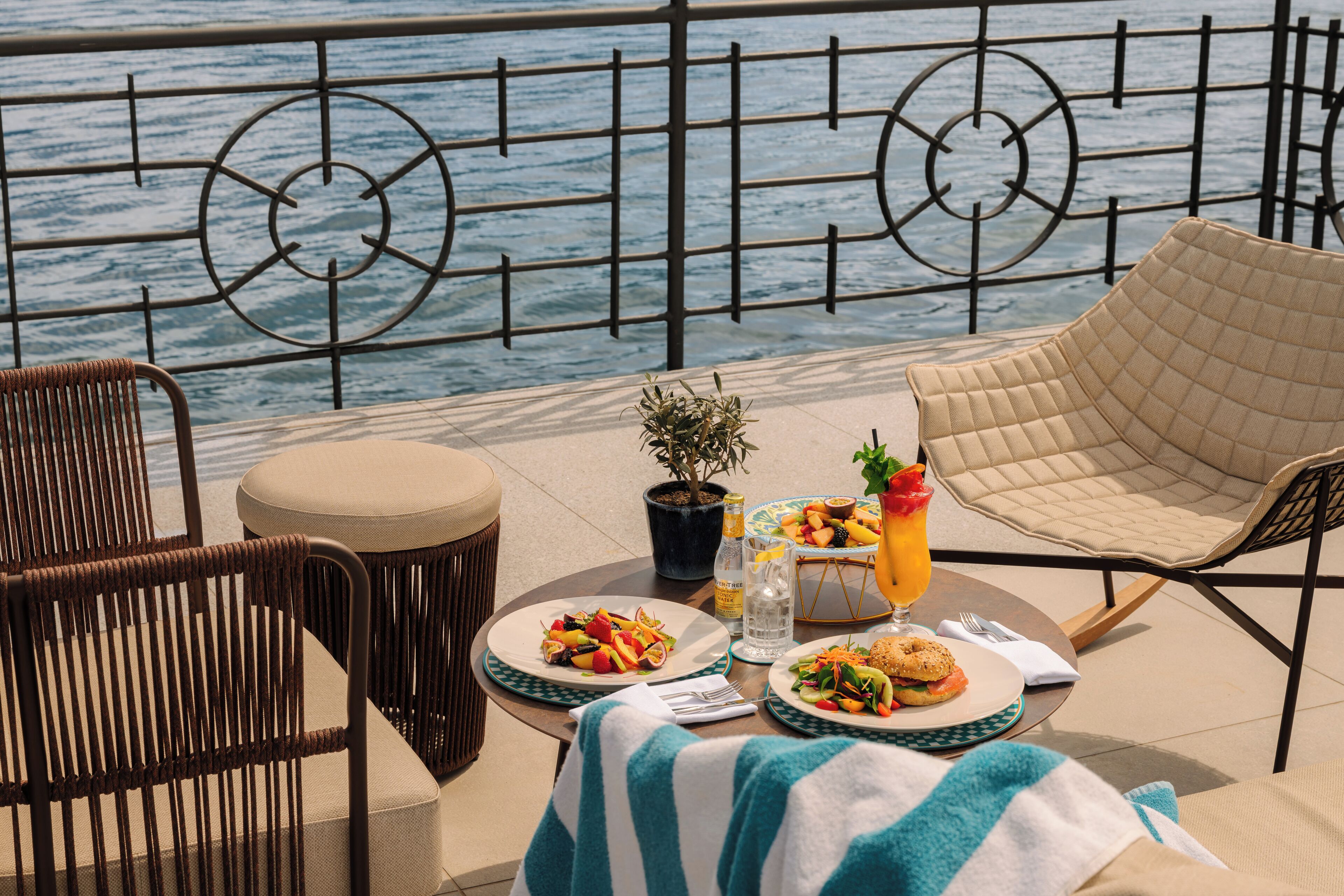 A dining area with a view of the sea, featuring wicker chairs, a round table with plates of food, a potted plant, and a striped towel.
