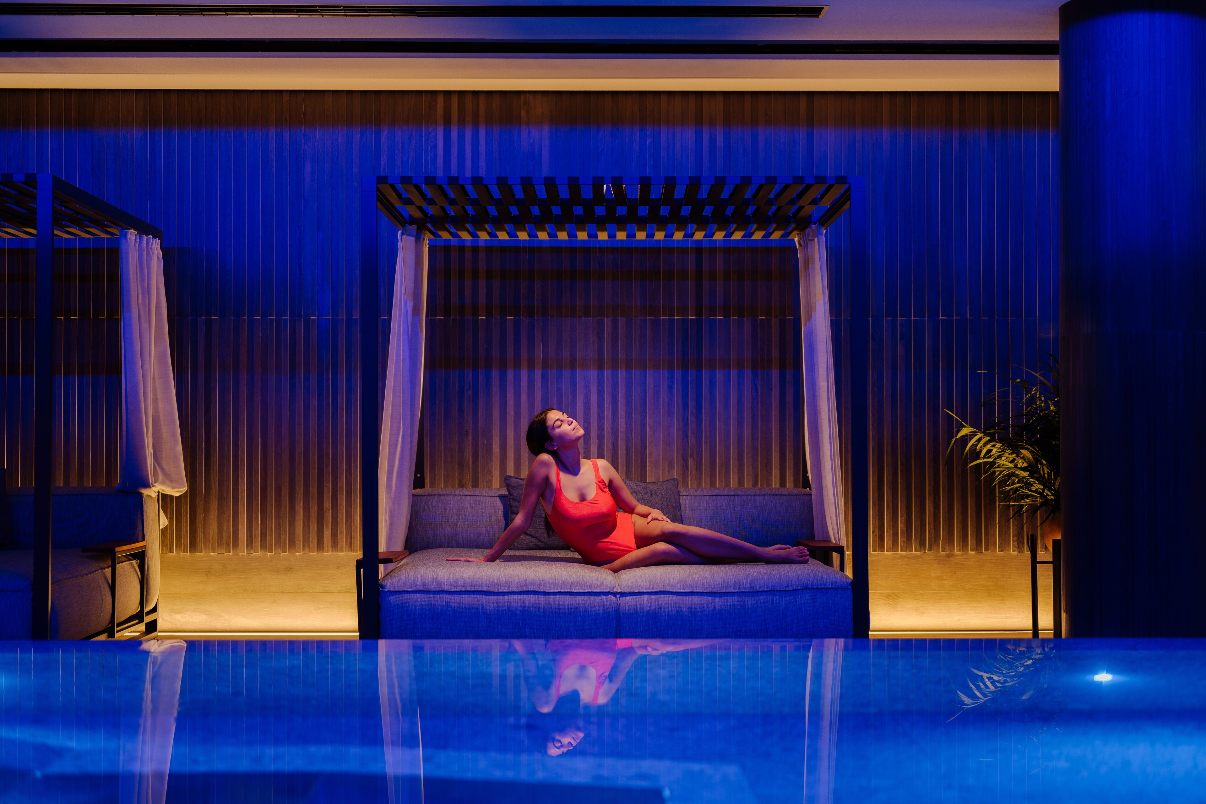 A woman in a red swimsuit sitting on a sofa by a pool with a blue-lit background.