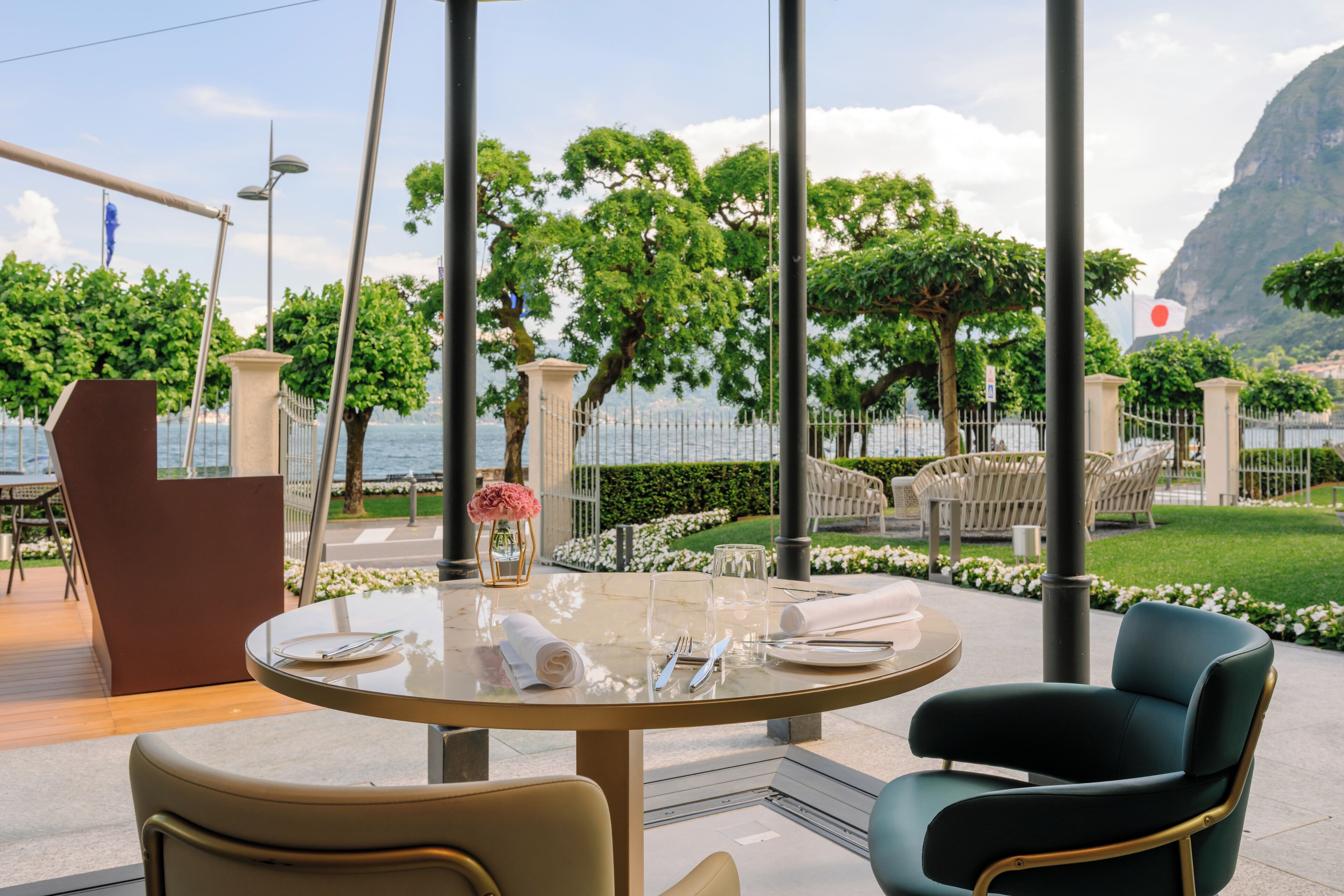 A round table with a glass top, surrounded by chairs, set outdoors with a view of a body of water and greenery in the background.