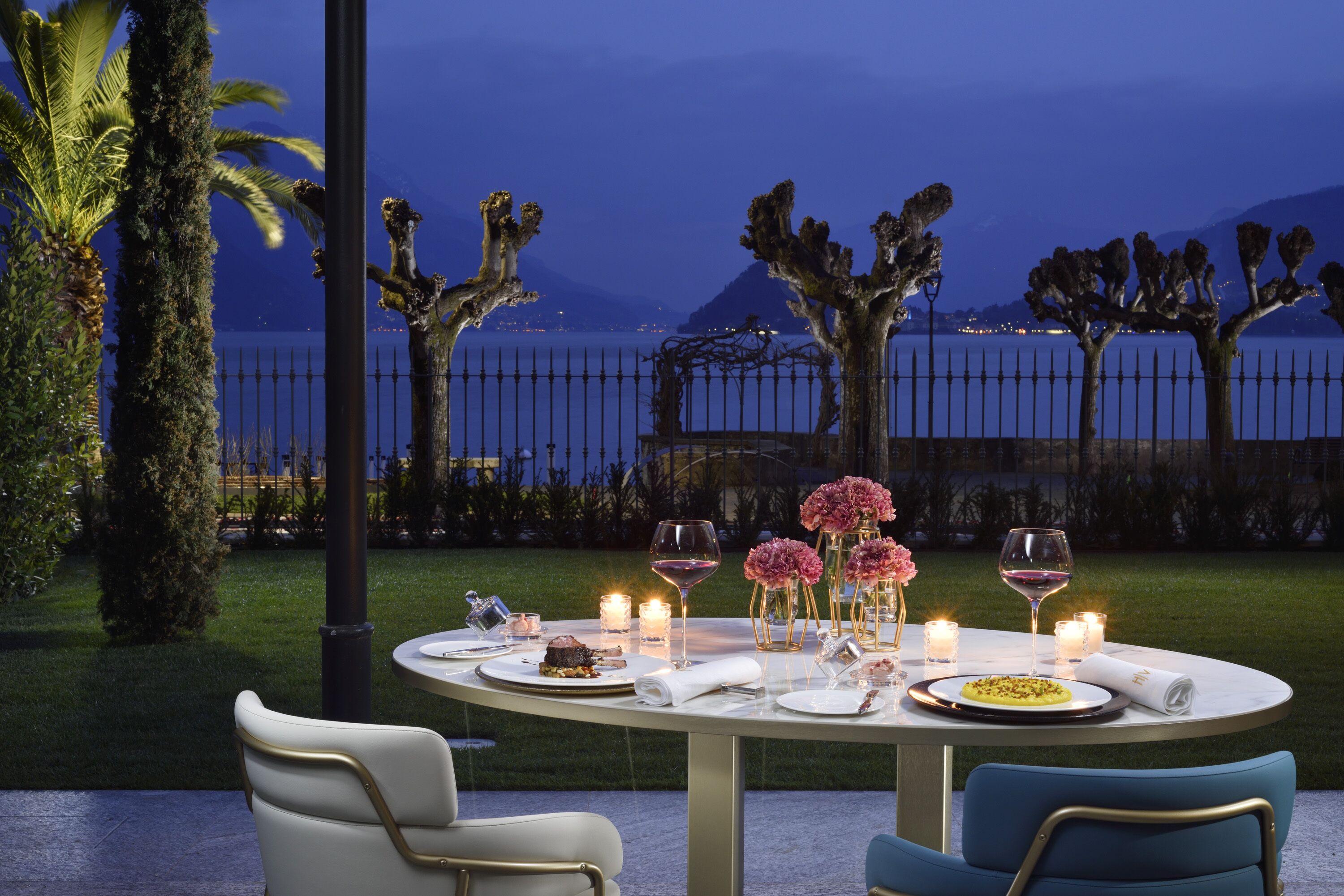 An outdoor dining area with a table set for two, surrounded by chairs, with a view of the sea and mountains in the background.