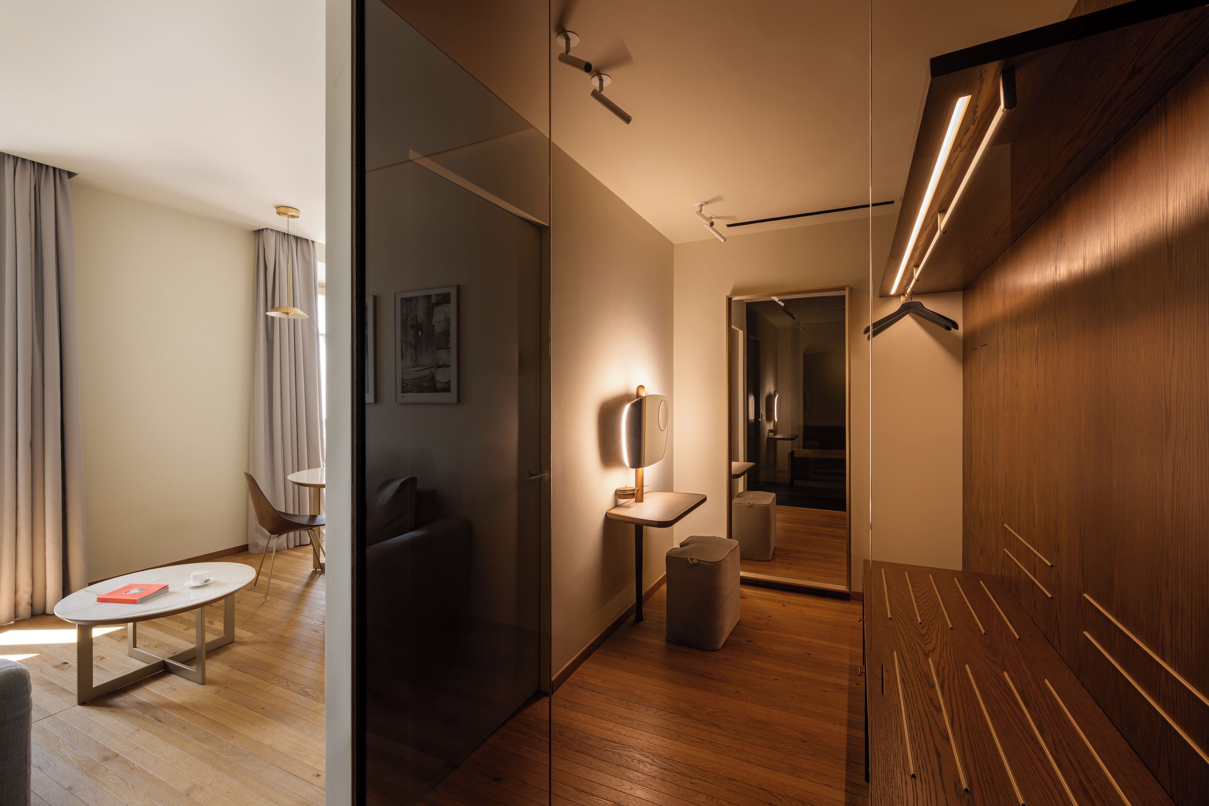 Et moderne hotelværelse med trægulv, et rundt bord med en rød bog, en stol, et natbord med en lampe og et garderobeskab med et spejl.
