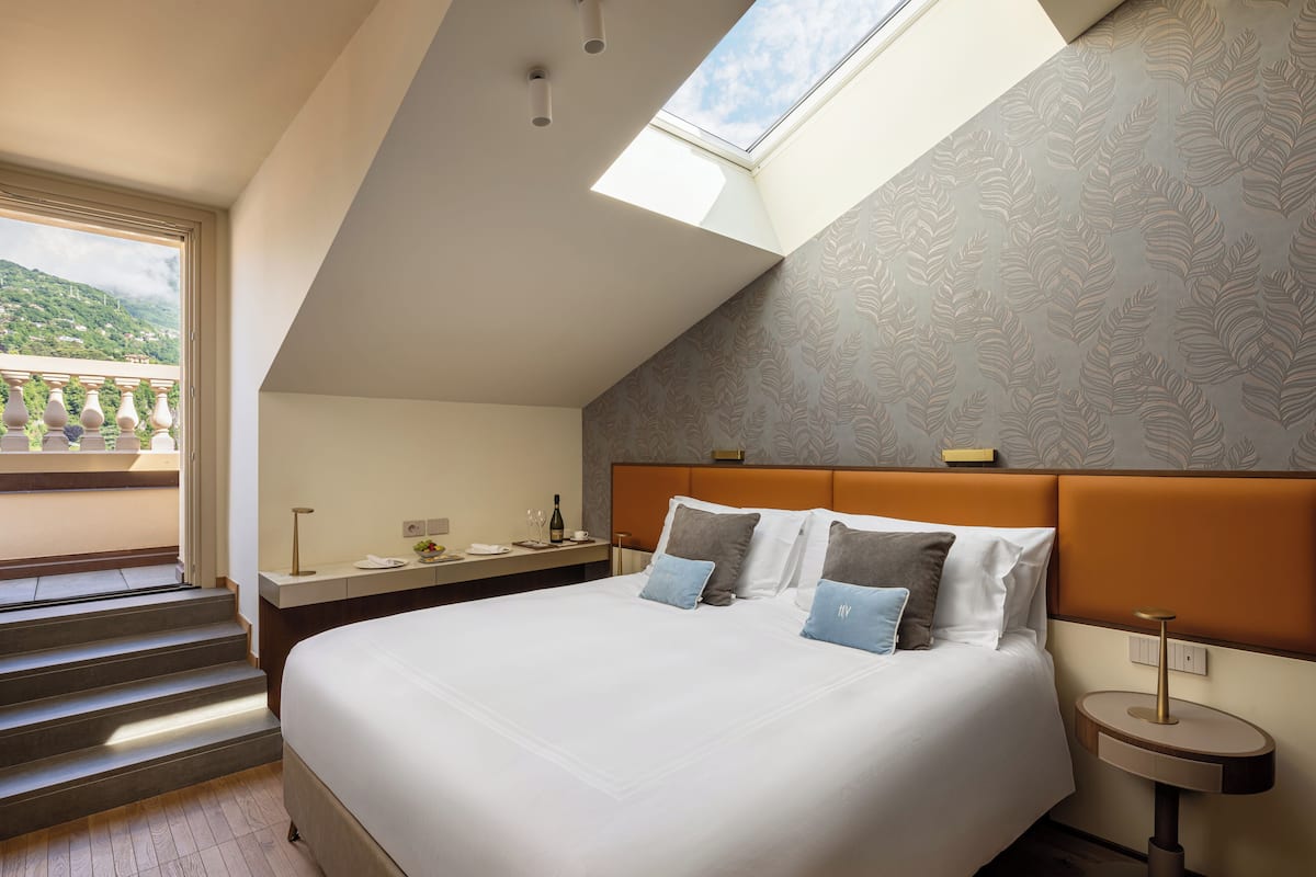 A modern hotel room with a large bed, a skylight, a view of the outdoors, and a small bedside table with a lamp.