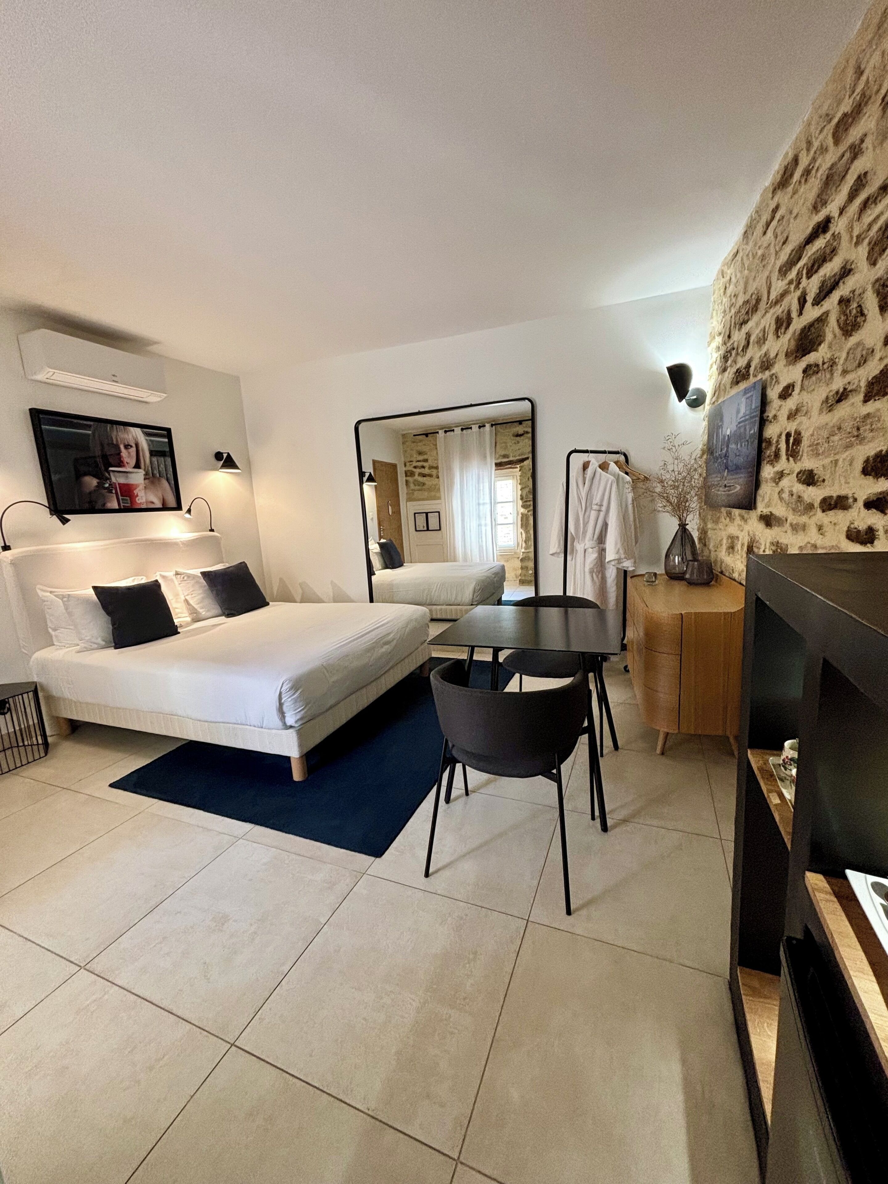 A modern hotel room with a large bed, a small table, a mirror, and a stone wall.