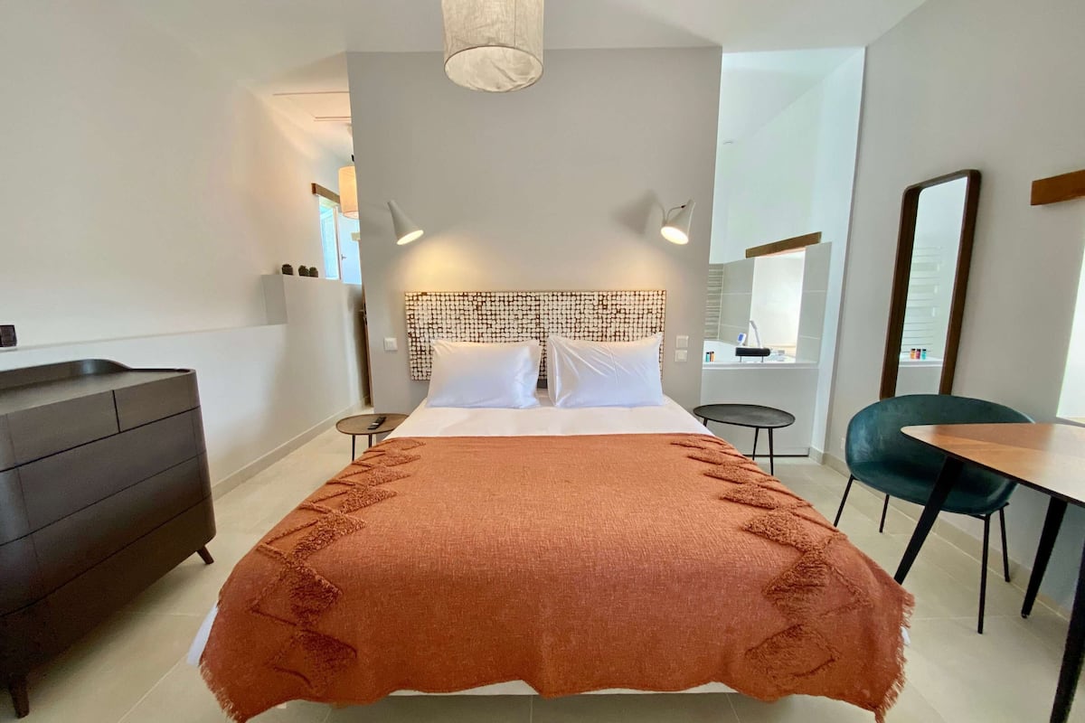 Ein modernes Hotelzimmer mit einem großen Bett, einer dunklen Holzkommode, einem kleinen runden Tisch, einem grünen Sessel und einem großen Spiegel.