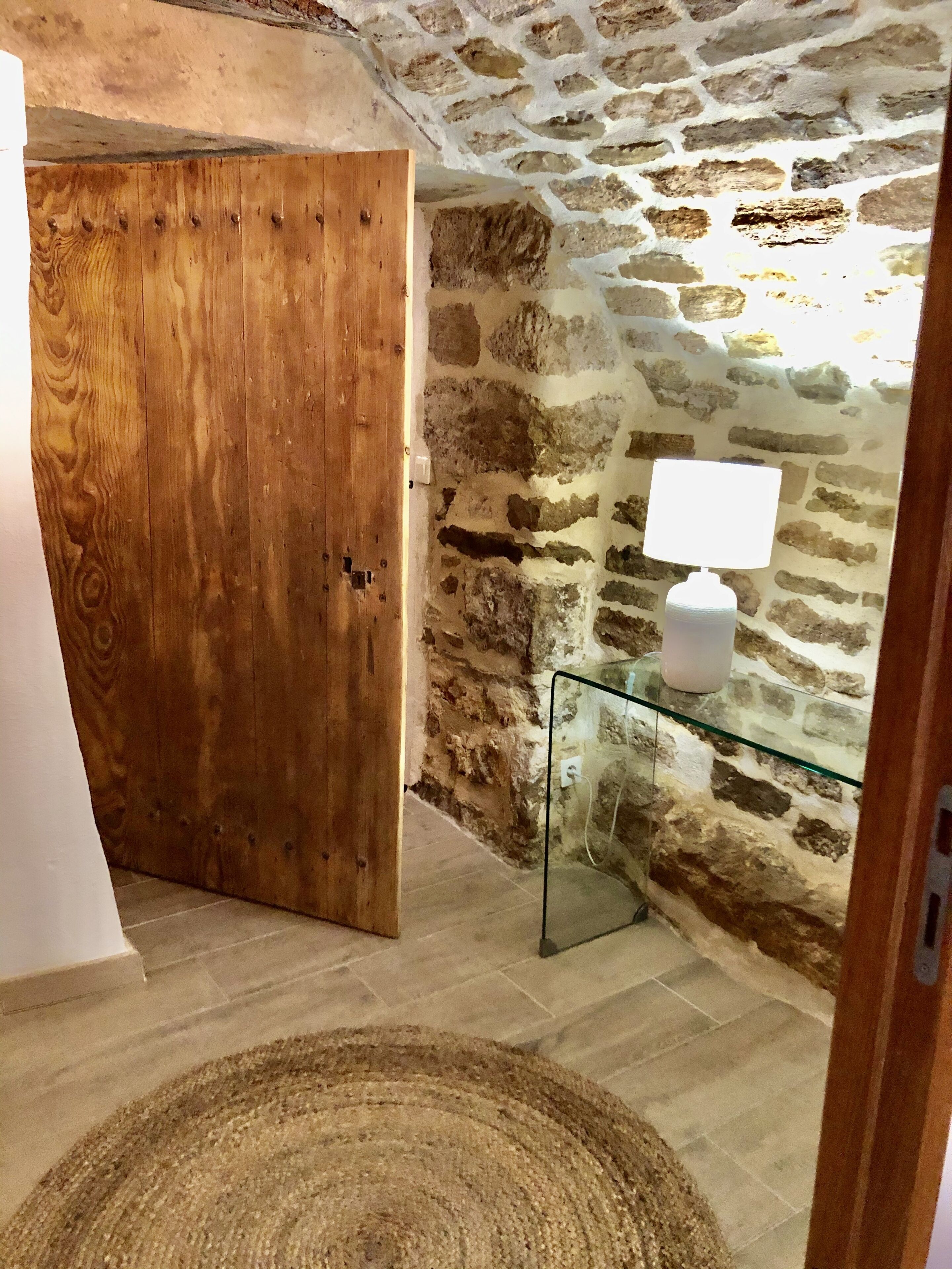 Una habitación con puerta de madera, una mesa de vidrio con una lámpara blanca y una pared de piedra.