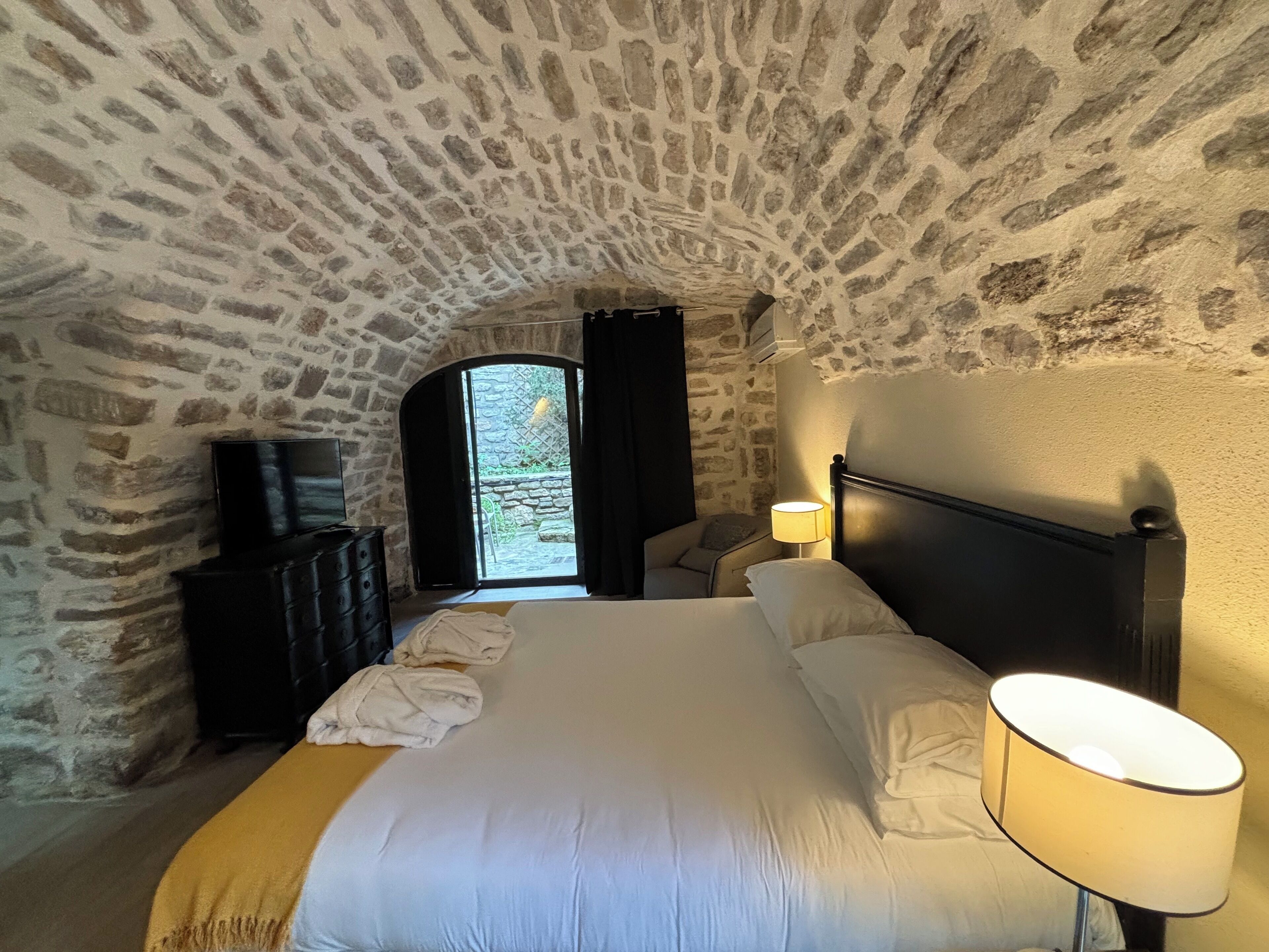 Un dormitorio con techo de piedra, una cama con ropa de cama blanca, una lámpara de noche y un televisor sobre un tocador.