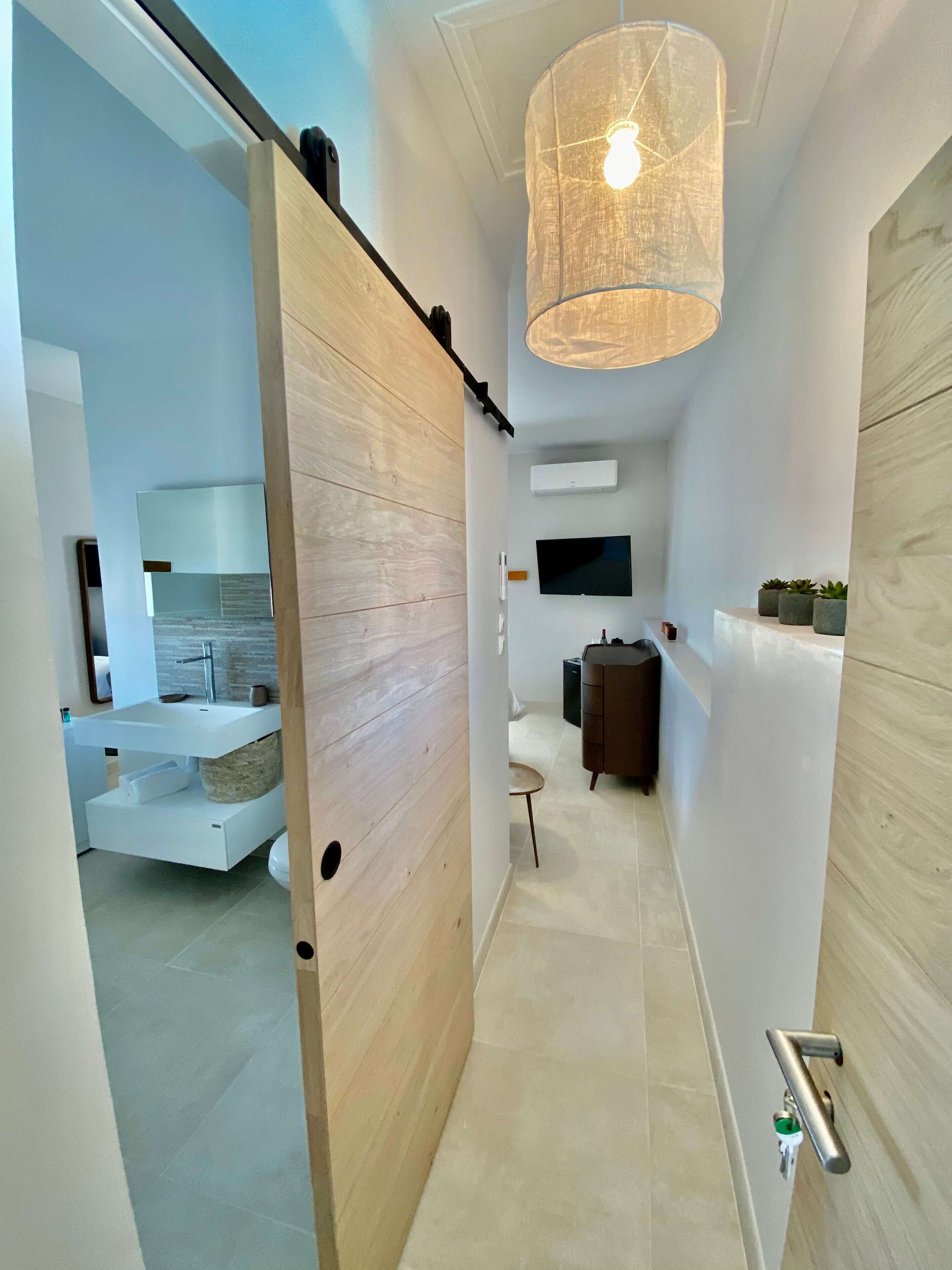 Un intérieur moderne avec une porte en bois, un luminaire suspendu, un téléviseur blanc fixé au mur et une petite table avec une chaise.