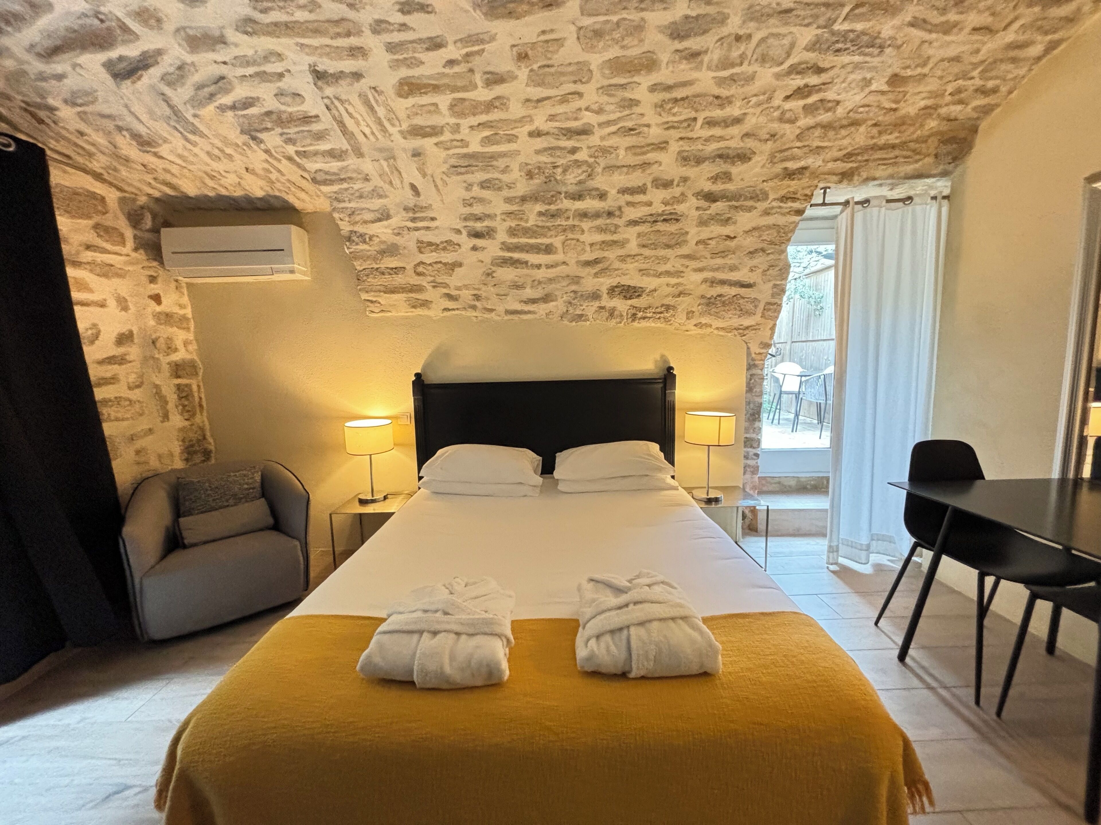A hotel room with a stone ceiling, a bed with two towels, a desk, and a chair.