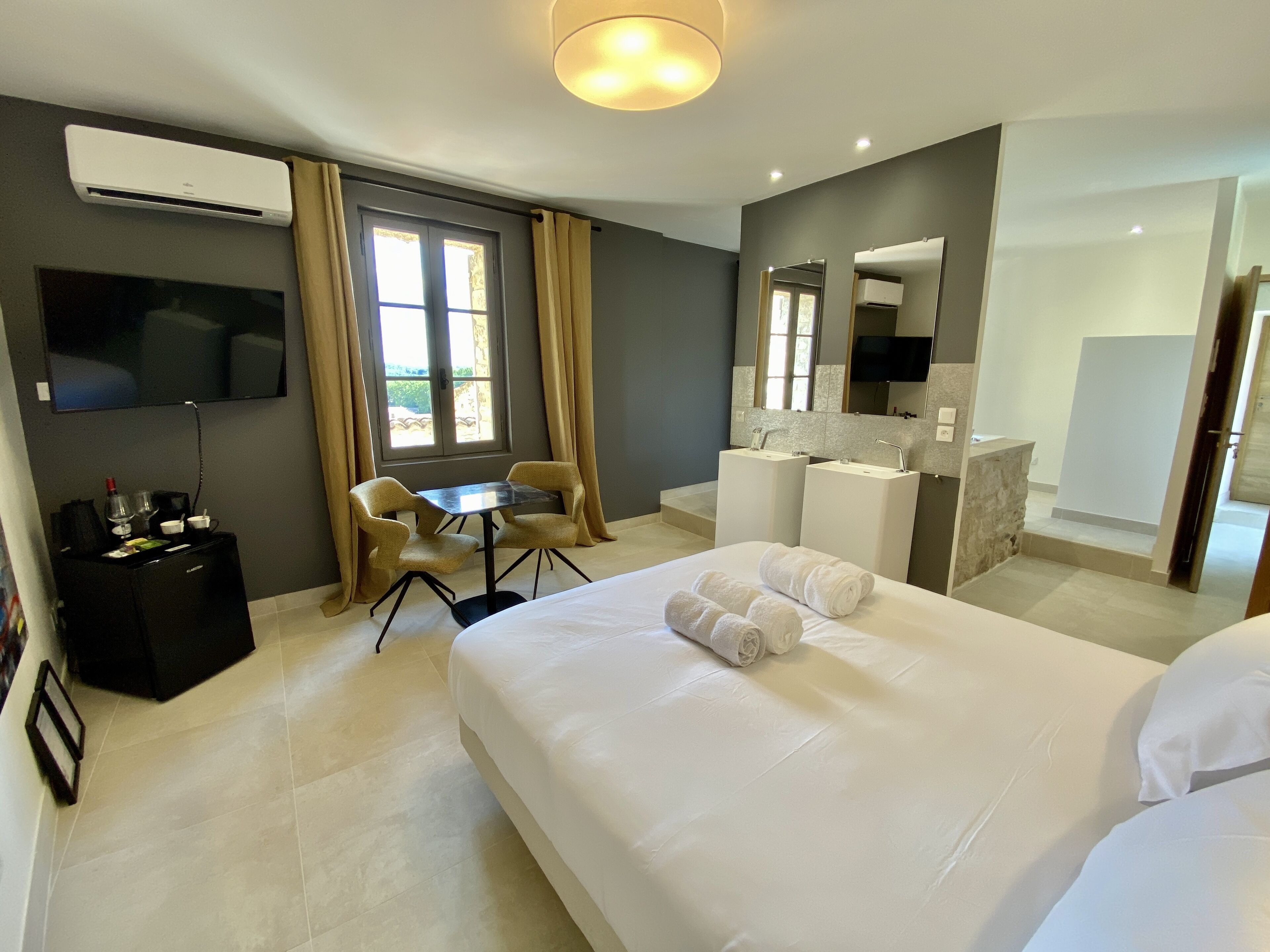 A hotel room with a bed, a desk, a chair, a TV, a minibar, and a bathroom with a mirror and sink.