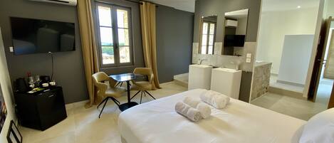 A hotel room with a bed, a desk, a chair, a TV, a minibar, and a bathroom with a mirror and sink.