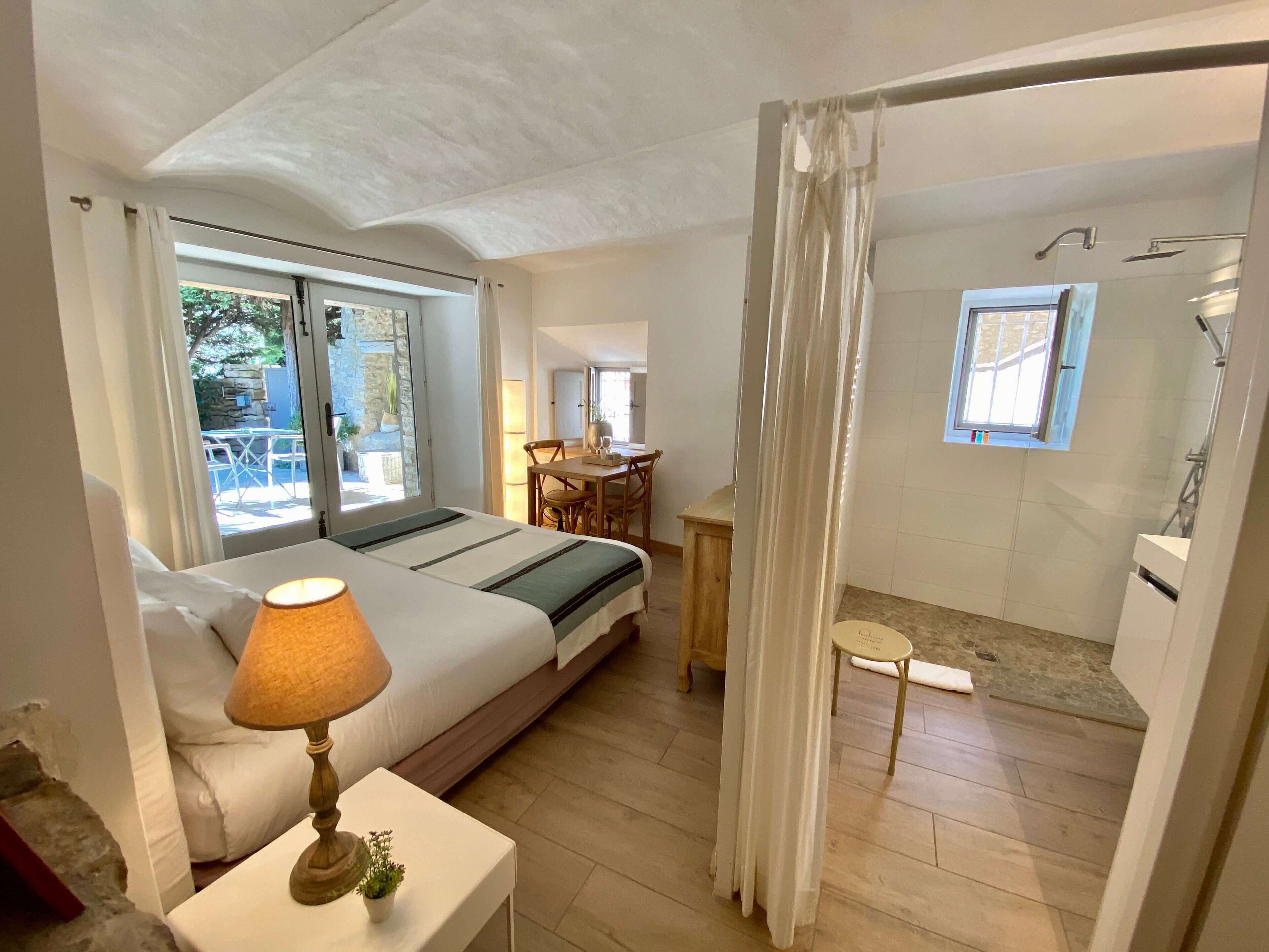 A bedroom with a bed, bedside lamp, and a view of the outside. There is a small table with a chair and a window.