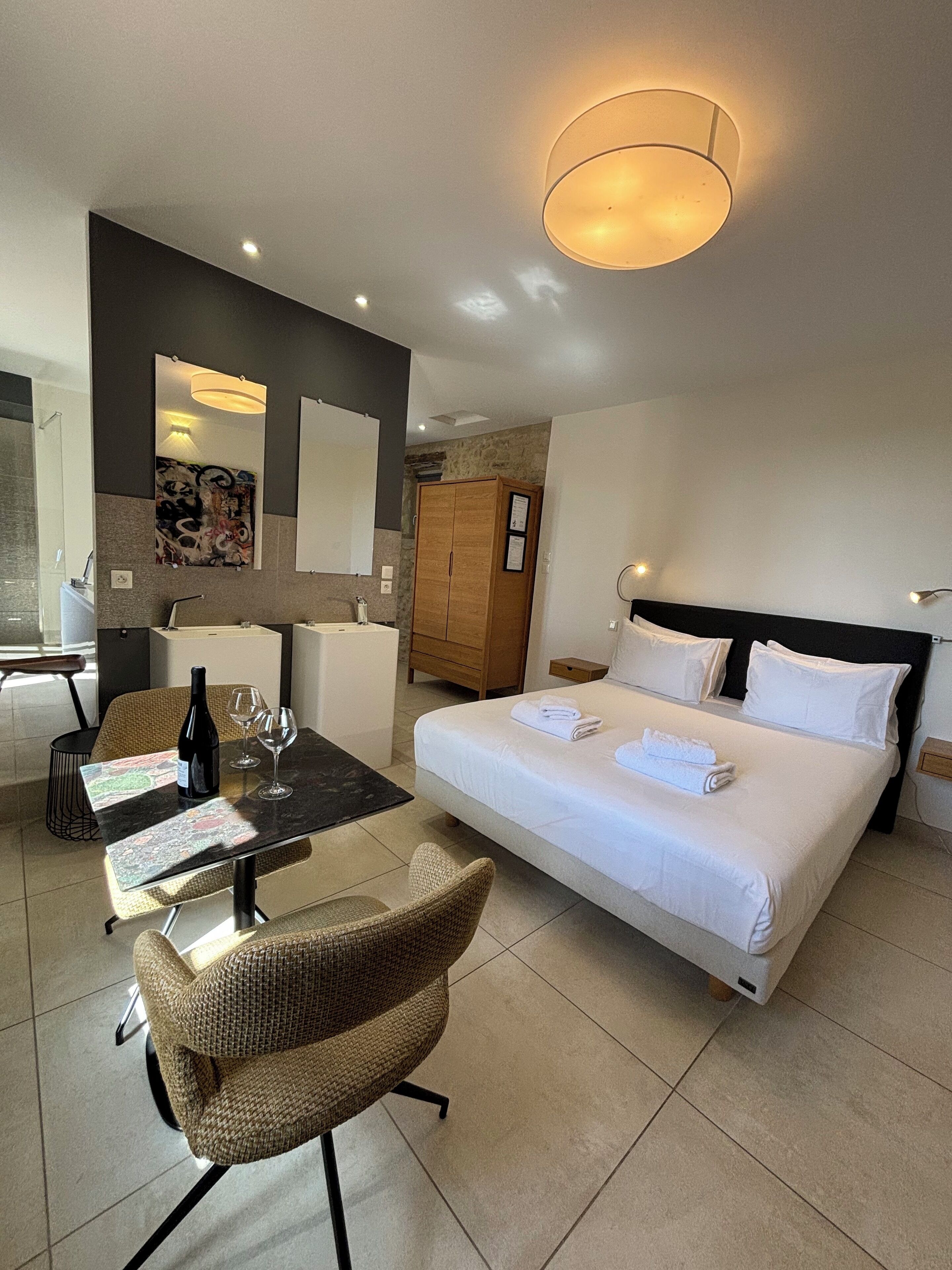 Ein Hotelzimmer mit einem Bett, einem Schreibtisch mit Stuhl, einem kleinen Tisch mit Flasche und Gläsern, einem Kleiderschrank und einem Spiegel.