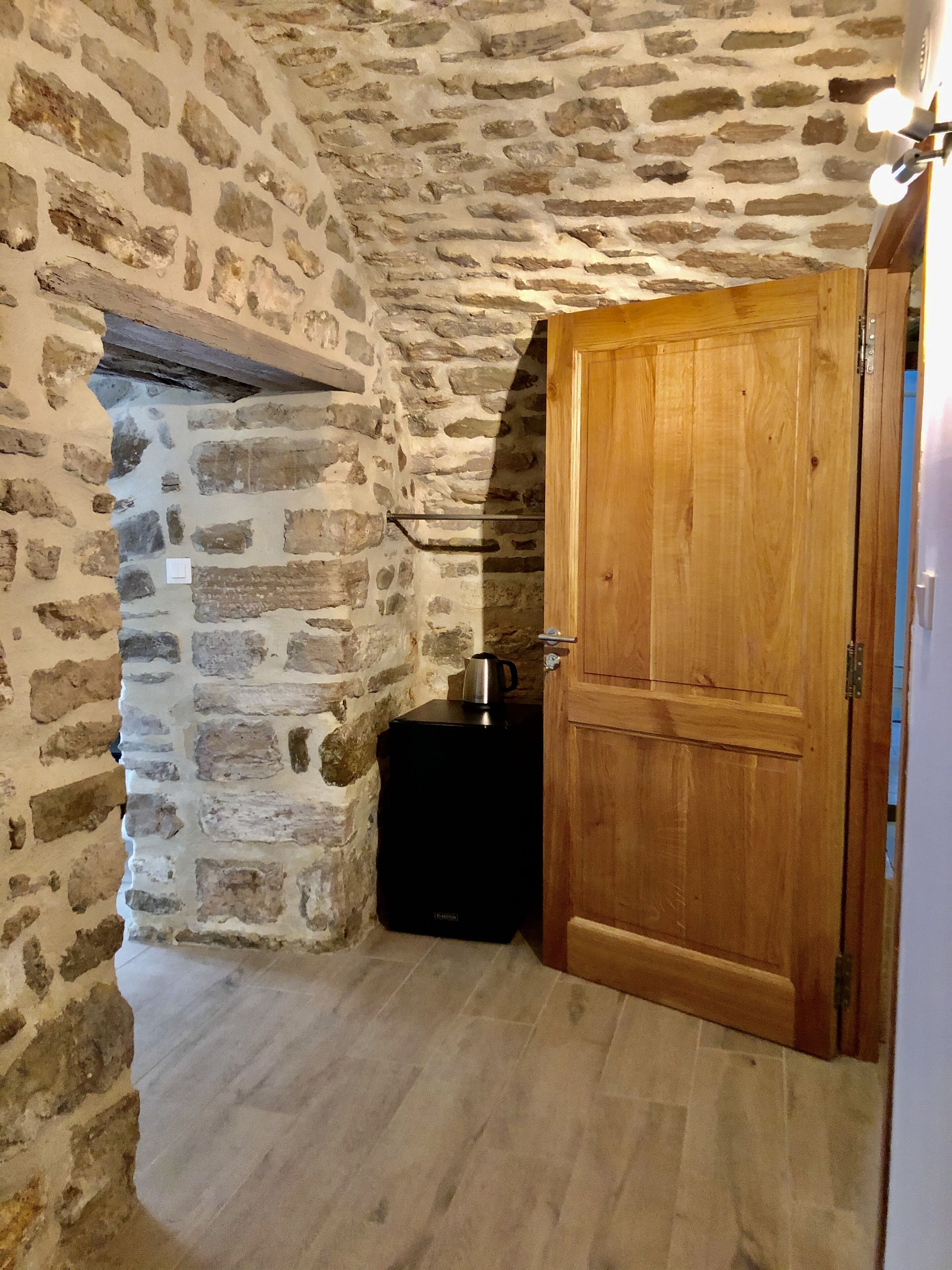 Ein Zimmer mit steinernen Wänden, einer Holztür, einem schwarzen Gerät und einem kleinen Regal.