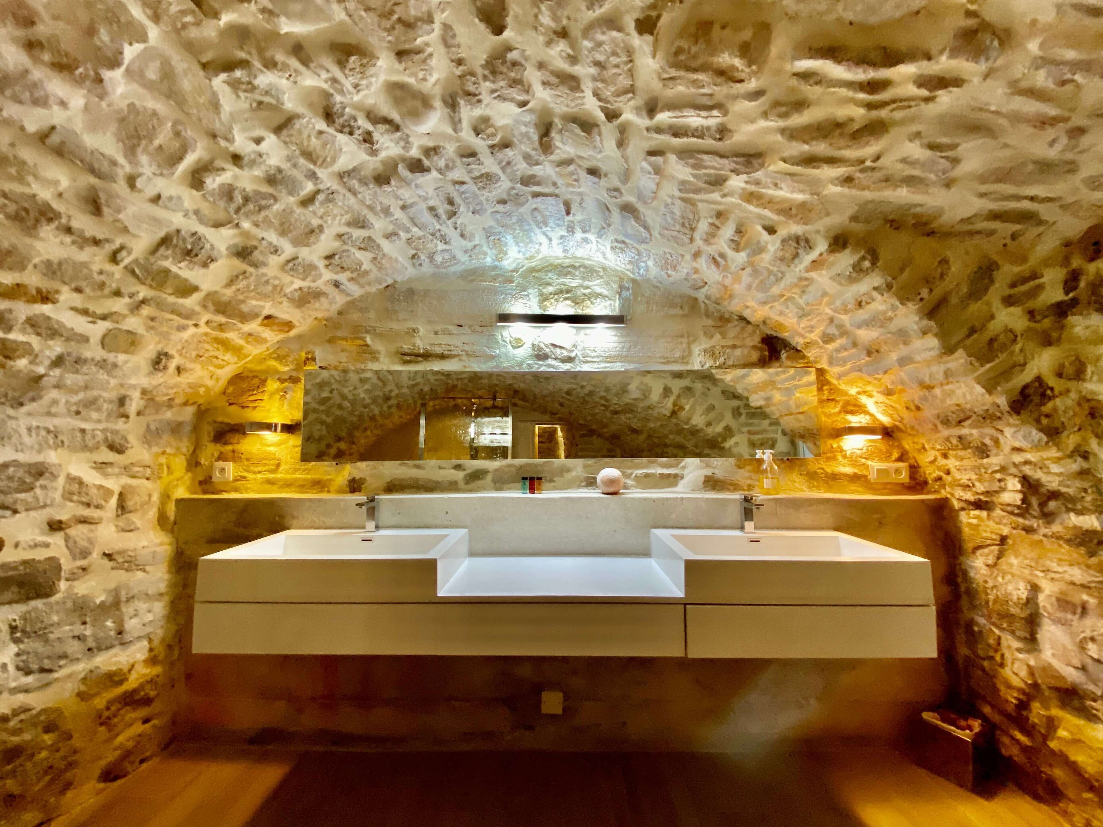 Un baño con paredes de piedra, un espejo grande, dos lavamanos y suelo de madera.
