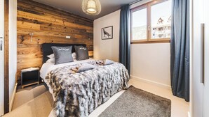 Una habitació amb paret de fusta, un llit amb una manta de pell, una tauleta de nit, una làmpada i una finestra amb cortines.