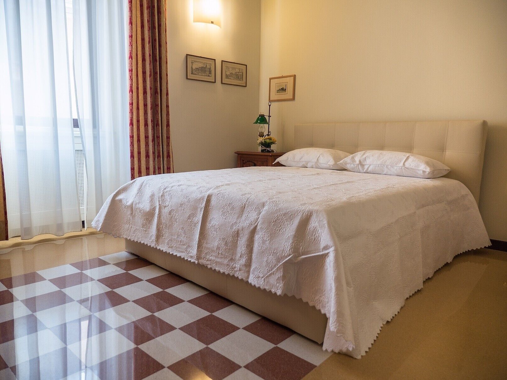 A hotel room with a large bed, a checkered floor, a nightstand with a lamp, and a window with curtains.