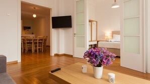 Ein modernes Wohnzimmer mit Holzboden, einem Wand-Fernseher, einem Essbereich mit Tisch und Stühlen sowie einem Bett mit Nachttisch.