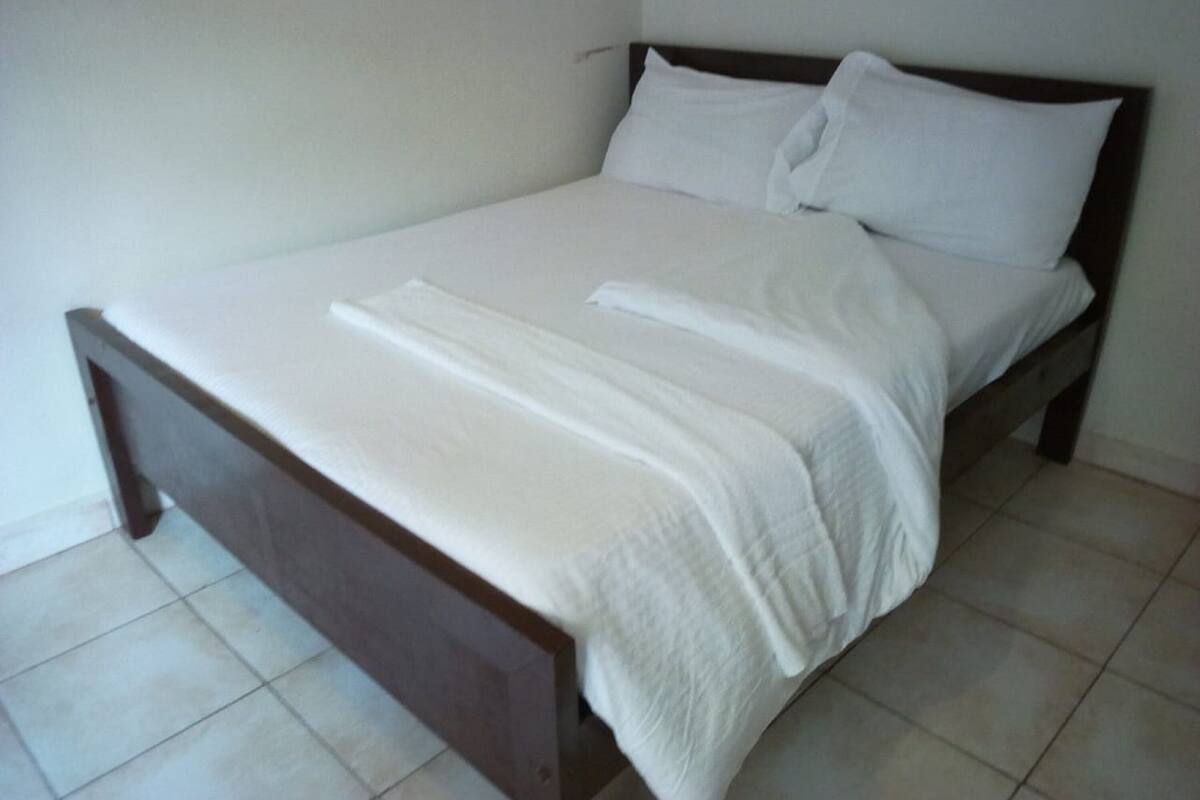 A single bed with a wooden frame and white bedding in a room with plain walls and tiled flooring.