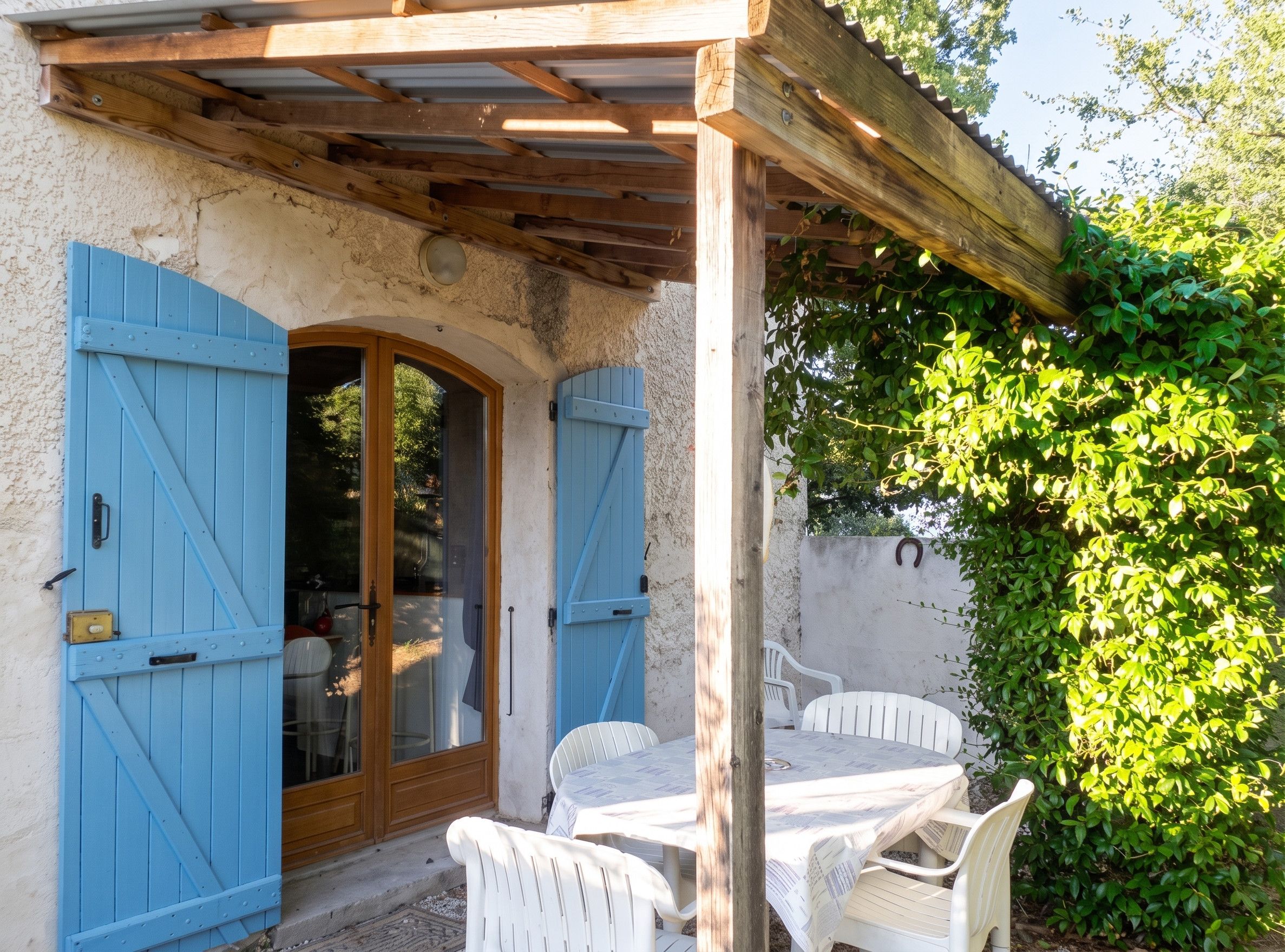 A covered outdoor seating area with a table and chairs, a wooden pergola, and a blue door with a glass panel.