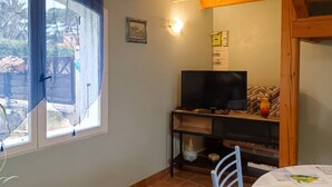 A room with a TV on a wooden stand, a small table with a chair, a window with curtains, and a wall-mounted light fixture. - Homerez - Charming Apt in Le Luc (Le Luc)