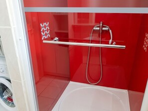 Une douche avec un flexible chromé et une porte en verre, une baignoire blanche et un mur recouvert de carreaux rouges.