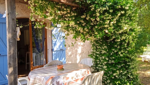 A rustic outdoor dining area with a table and chairs, a wooden pergola, and a lush green bush with white flowers. - Homerez - Charming Apt in Le Luc (Le Luc)