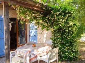 Un espace de restauration extérieur rustique, avec une table et des chaises, une pergola en bois et un arbuste luxuriant aux feuilles vertes et aux fleurs blanches.